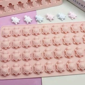 May include: A pink silicone mould featuring turtle shapes. The mould has multiple turtle impressions, and there are several small, colourful turtle-shaped sweets in white, blue, and pink. The mould is designed for making sweets or other small treats.