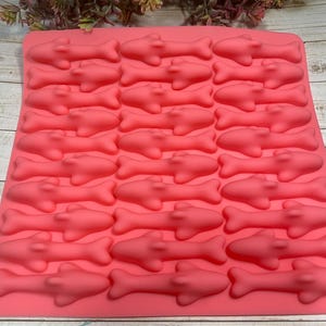 May include: A coral-colored silicone mold with a repeating pattern of shark shapes. The mold is rectangular and features multiple shark cavities, perfect for creating themed treats or crafts. The sharks are detailed with fins and a streamlined body.