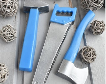 Op de afbeelding: Drie speelgoedgereedschappen gemaakt van plastic en metaal. Een blauwe hamer, een blauwe zaag met een zilveren blad en een blauwe bijl met een zilveren blad.