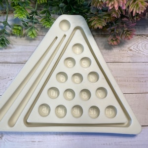 May include: White silicone mold shaped like a triangle with a pool table design. The mold has 15 round indentations for making billiard balls, numbered 1 through 15. There are also two long indentations for making pool cues.