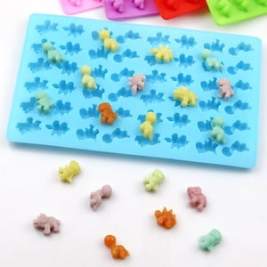 May include: A blue silicone mold with 36 dinosaur-shaped cavities.  The mold is filled with colorful dinosaur-shaped candies.  There are also several dinosaur-shaped candies scattered on a white surface.