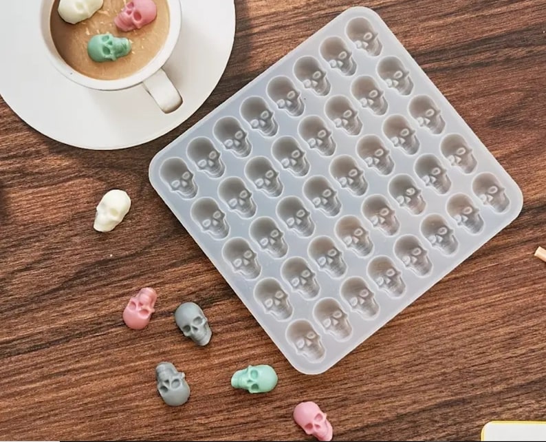 May include: A clear silicone mold with 42 skull-shaped cavities. The mold is on a wooden surface with several small, colorful skull-shaped candies.