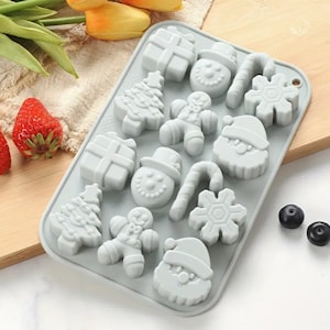 May include: A light gray silicone mold with Christmas-themed shapes, including a Christmas tree, snowman, Santa Claus, gingerbread man, snowflake, candy cane, and gift box. The mold is designed for making festive treats.