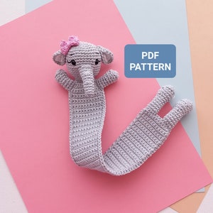 Pode incluir: Um marcador de livro de elefante de crochê cinza com um laço rosa na cabeça. O marcador está sobre um fundo rosa com um fundo azul e pêssego atrás. O texto "PDF PATTERN" está em um retângulo azul.