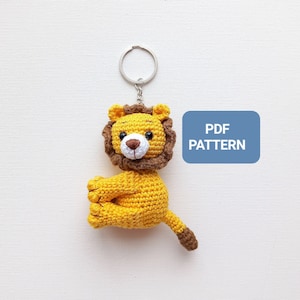 Op de afbeelding: Een gehaakte leeuwen sleutelhanger met een bruine manen, een geel lichaam en een wit gezicht. De sleutelhanger is bevestigd aan een zilveren ring. De tekst "PDF PATTERN" staat op een blauwe rechthoek.