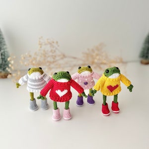 May include: Four crocheted frog figurines wearing sweaters and boots. The frogs are wearing a grey, red, pink, and yellow sweater. The sweaters have white hearts on them. The frogs are standing on a white surface.