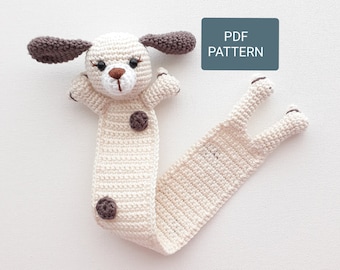 May include: Crocheted dog bookmark with brown ears and spots. The bookmark is white with a long tail. The text "PDF PATTERN" is on a blue rectangle.