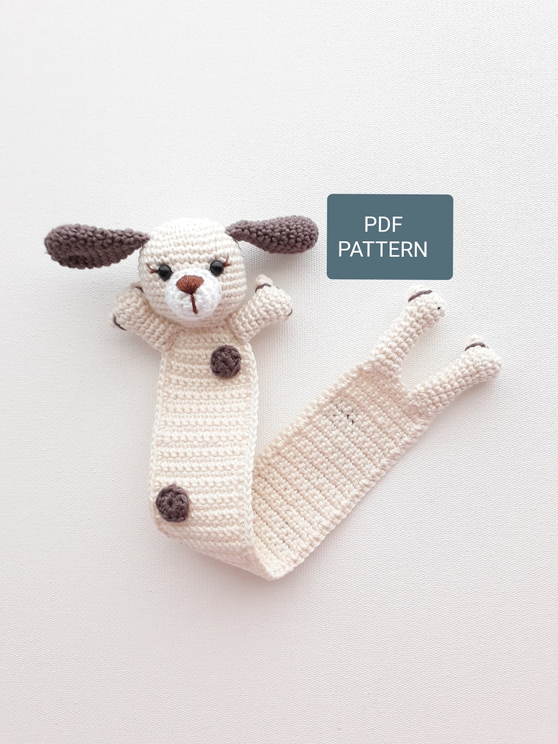 Puede incluir: Marcap&aacute;ginas de crochet con forma de perro con orejas y manchas marrones. El marcap&aacute;ginas es blanco con una cola larga. El texto "PDF PATTERN" est&aacute; en un rect&aacute;ngulo azul.