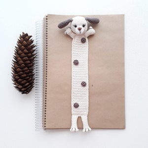 Puede incluir: Un marcap&aacute;ginas de crochet con forma de perro con manchas marrones sobre fondo blanco. El marcap&aacute;ginas est&aacute; sobre una libreta marr&oacute;n.