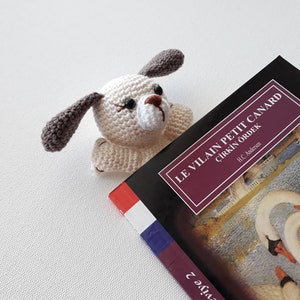 Puede incluir: Un marcap&aacute;ginas de crochet blanco y marr&oacute;n con forma de perro sobre un libro titulado "Le Vilain Petit Canard" con el subt&iacute;tulo "&Ccedil;irkin &Ouml;rdek" de H.C. Andersen. La portada del libro muestra una pintura de dos cisnes blancos.