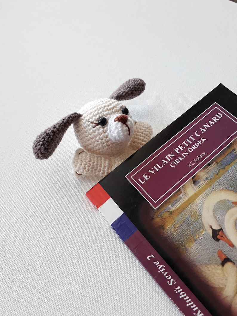 Puede incluir: Un marcap&aacute;ginas de crochet blanco y marr&oacute;n con forma de perro sobre un libro titulado "Le Vilain Petit Canard" con el subt&iacute;tulo "&Ccedil;irkin &Ouml;rdek" de H.C. Andersen. La portada del libro muestra una pintura de dos cisnes blancos.