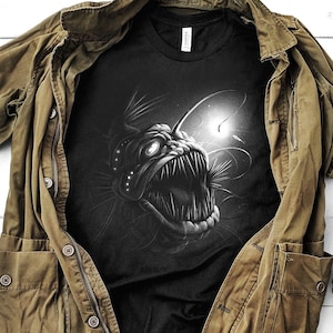K&ouml;nnte beinhalten: Ein schwarzes T-Shirt mit einem grafischen Aufdruck eines Anglerfisches. Das T-Shirt liegt auf einer wei&szlig;en Holzoberfl&auml;che mit einer braunen Jacke, zwei braunen Stiefeln und drei kleinen gr&uuml;nen Pflanzen.
