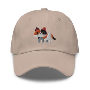 Può includere: Un cappello da baseball beige con un design di gatto calico ricamato. Il gatto è grigio, bianco e arancione con occhi neri.