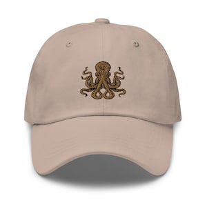 Könnte beinhalten: Eine beige Baseballkappe mit einem gestickten braunen Oktopus-Design.