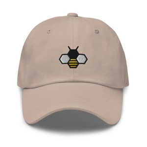 Op de afbeelding: Een beige baseballpet met een geborduurd bijendesign. De bij is zwart en wit met gele strepen.