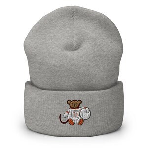May include: Gray knitted beanie with a cuffed brim. The beanie features an embroidered design of a brown monkey wearing an orange and white astronaut suit holding a white helmet.