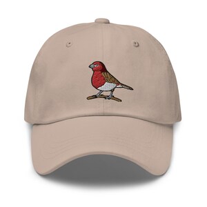 Könnte beinhalten: Eine beige Baseballkappe mit einem gestickten roten und braunen Vogel auf der Vorderseite.