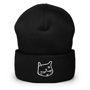 May include: Black beanie with a white embroidered cat face. The cat has a grumpy expression and a small X over its eye.