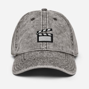 May include: A gray distressed denim baseball cap with a black and white embroidered clapperboard design.