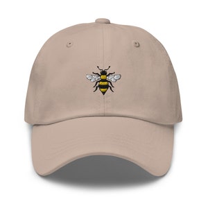 Op de afbeelding: Een beige baseballpet met een geborduurd bijendesign. De bij is zwart en geel met witte vleugels.