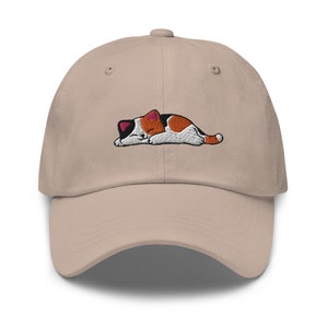 Può includere: Un cappello da baseball beige con un'immagine ricamata di un gatto calico che dorme. Il gatto è arancione, bianco e nero.