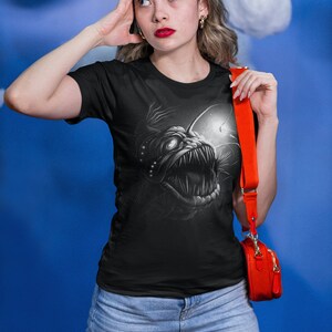 K&ouml;nnte beinhalten: Ein schwarzes T-Shirt mit einem grafischen Aufdruck eines Tiefsee-Anglerfisches. Der Anglerfisch ist in einem wei&szlig;en und grauen Farbschema dargestellt. Das T-Shirt wird von einer Person mit einer roten Umh&auml;ngetasche getragen.