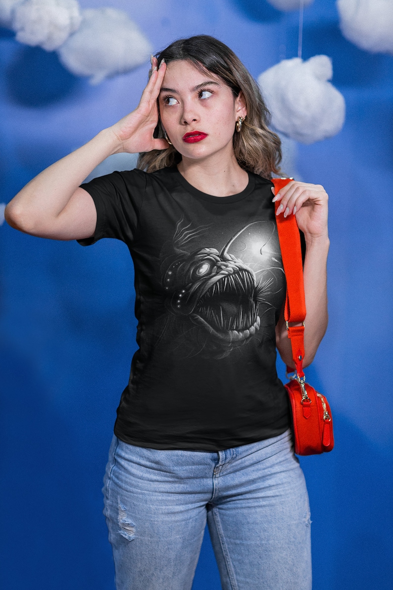 K&ouml;nnte beinhalten: Ein schwarzes T-Shirt mit einem grafischen Aufdruck eines Tiefsee-Anglerfisches. Der Anglerfisch ist in einem wei&szlig;en und grauen Farbschema dargestellt. Das T-Shirt wird von einer Person mit einer roten Umh&auml;ngetasche getragen.