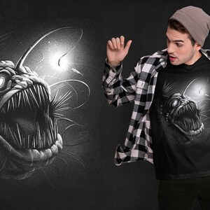 K&ouml;nnte beinhalten: Ein schwarzes T-Shirt mit einem wei&szlig;en Grafikdruck eines Tiefsee-Anglerfisches mit einer biolumineszierenden K&ouml;der. Der Anglerfisch ist mit scharfen Z&auml;hnen und einem bedrohlichen Ausdruck dargestellt.