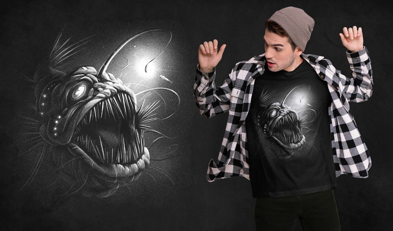 K&ouml;nnte beinhalten: Ein schwarzes T-Shirt mit einem wei&szlig;en Grafikdruck eines Tiefsee-Anglerfisches mit einer biolumineszierenden K&ouml;der. Der Anglerfisch ist mit scharfen Z&auml;hnen und einem bedrohlichen Ausdruck dargestellt.