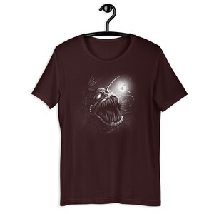 K&ouml;nnte beinhalten: Ein maroonfarbenes T-Shirt mit einer wei&szlig;en Grafik eines Tiefsee-Anglerfisches mit einem biolumineszierenden K&ouml;der.