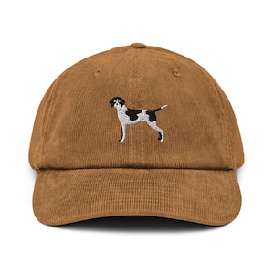 May include: A brown corduroy baseball cap with an embroidered black and white dog design. The cap has a curved peak and an adjustable strap at the back. The dog illustration is detailed and centered on the front of the cap.