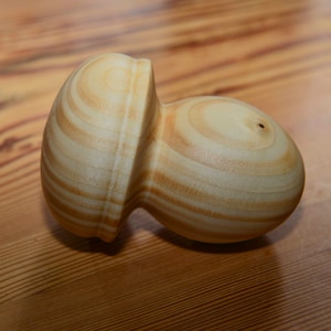 Puede incluir: Un tapón de botella de madera clara con una forma orgánica única. El tapón tiene una parte superior redondeada y una base bulbosa, con patrones de vetas de madera visibles. El artículo es probablemente hecho a mano y diseñado para uso decorativo.