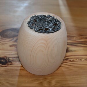 Könnte beinhalten: Ein Holzbehälter mit einem dekorativen Metalldeckel. Der Deckel hat ein florales Design mit einem Vogel in der Mitte.