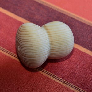 Puede incluir: Un objeto de madera con dos secciones redondeadas y bulbosas, cada una con una textura acanalada. El objeto es de color marrón claro y descansa sobre una superficie rayada roja y naranja.