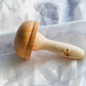 Może przedstawiać: Jasne, drewniane narzędzie do masażu z główką w kształcie grzyba i długim, zaokrąglonym uchwytem. Główka ma żebrowany wzór, a uchwyt gładkie, naturalne wykończenie. Przeznaczone do masażu i relaksu.