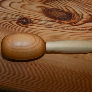 Puede incluir: Un mazo de madera con un mango de color claro y una cabeza redondeada. La cabeza es de color marrón claro y el mango es de un tono de madera natural más claro. El mazo está sobre una superficie de madera.