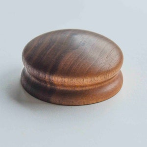 Puede incluir: Un molinillo de madera artesanal con un acabado marrón oscuro. El molinillo tiene una parte superior redondeada y una base ligeramente más ancha, mostrando la veta natural de la madera. Diseño sencillo y funcional.