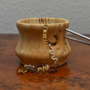 Puede incluir: Un cuenco de hilo de madera con un acabado natural, con un diseño curvo y una ranura para el hilo. Dos agujas de tejer plateadas están insertadas en el cuenco. Se ven hebras de hilo en tonos marrón, amarillo y blanco.