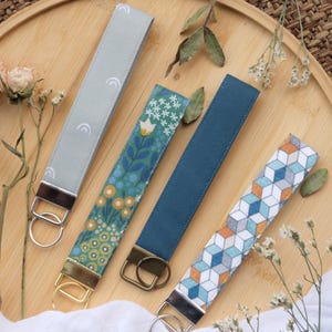 May include: Four fabric wristlet keychains with metal hardware. One is light grey with white arches, one features a green floral pattern, one is solid dark blue, and one has a geometric cube pattern in blue, orange, and white. Each has a metal ring.