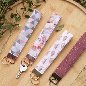 May include: Four fabric wristlet keychains with metal hardware. One features a geometric triangle pattern, one a floral print, one a rainbow motif on white, and one a mauve fabric with white polka dots. One keychain includes a silver metal key.