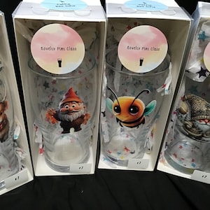 May include: Four novelty pint glasses featuring different cartoon designs. The glasses are packaged in clear boxes with a white background and a pink circle with the text "Novelty Pint Glass". The designs include a Highland cow, a gnome, a bee, and a snowman.