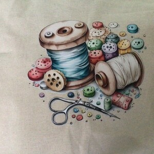 May include: A beige fabric surface featuring a printed illustration of two wooden spools of thread, one wound with light blue thread and the other with white thread, surrounded by a collection of colourful buttons and a pair of metal scissors.