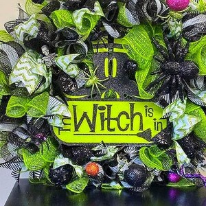 May include: A Halloween wreath with a black and neon green color scheme. The wreath features a sign that reads "The Witch is in". Decorative elements include spiders, glittery balls, and a skeleton.