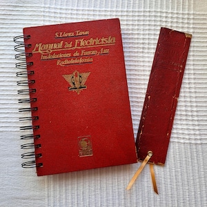 May include: A red spiral-bound book and matching bookmark. The book cover features gold lettering and an emblem. The bookmark has a gold ribbon. The book's title is "Manual del Electricista".