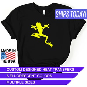 May include: Black t-shirt with a yellow frog silhouette graphic. The text "SHIPS TODAY!" is in a blue box at the top of the image. The text "MADE IN THE USA" is in a red, white, and blue box at the bottom left of the image. The text "CUSTOM DESIGNED HEAT TRANSFERS" is in a purple box at the bottom of the image. The text "6 FLUORESCENT COLORS" is in a purple box below the previous text. The text "MULTIPLE SIZES" is in a purple box below the previous text.