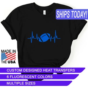 May include: Black t-shirt with a blue football and a heartbeat graphic. The text "SHIPS TODAY!" is in a white box with a blue border. The text "MADE IN THE USA" is in a red, white, and blue box. The text "CUSTOM DESIGNED HEAT TRANSFERS", "6 FLUORESCENT COLORS", and "MULTIPLE SIZES" are in white boxes with a purple border.