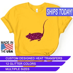 May include: Mustard yellow t-shirt featuring a glittery magenta mouse design. The shirt includes the text "MADE IN THE USA" and "SHIPS TODAY!". Additional text includes "CUSTOM DESIGNED HEAT TRANSFERS", "12 GLITTER COLORS", and "MULTIPLE SIZES".