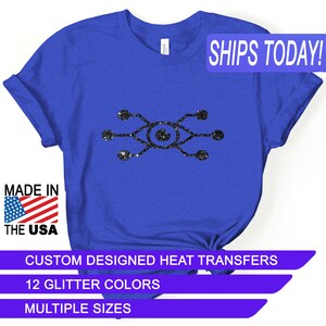 May include: A royal blue t-shirt with a black glitter design of an eye with lines extending from it. The text "SHIPS TODAY!" is in a white rectangle with a blue border. The text "MADE IN THE USA" is in a red, white, and blue rectangle. The text "CUSTOM DESIGNED HEAT TRANSFERS" is in a white rectangle with a purple border. The text "12 GLITTER COLORS" is in a white rectangle with a purple border. The text "MULTIPLE SIZES" is in a white rectangle with a purple border.