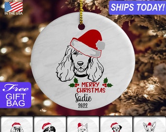 May include: White ceramic Christmas ornament with a black and white illustration of a dog wearing a red Santa hat. The ornament has the text "Merry Christmas Sadie 2022" on it.