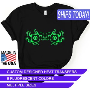 May include: Black t-shirt with a green graphic of two eyes with tentacles. The text "SHIPS TODAY!" is in a blue box at the top of the image. The text "MADE IN THE USA" is in a red, white, and blue box at the bottom left of the image. The text "CUSTOM DESIGNED HEAT TRANSFERS" is in a purple box at the bottom of the image. The text "6 FLUORESCENT COLORS" is in a purple box below the previous text. The text "MULTIPLE SIZES" is in a purple box below the previous text.